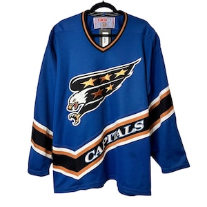 Puede incluir: Jersey de hockey azul con el logo de los Capitals de Washington en la parte delantera. El logo presenta un águila estilizada con estrellas y la palabra "CAPITALS". El jersey tiene cuello en V y detalles en negro, blanco y naranja.