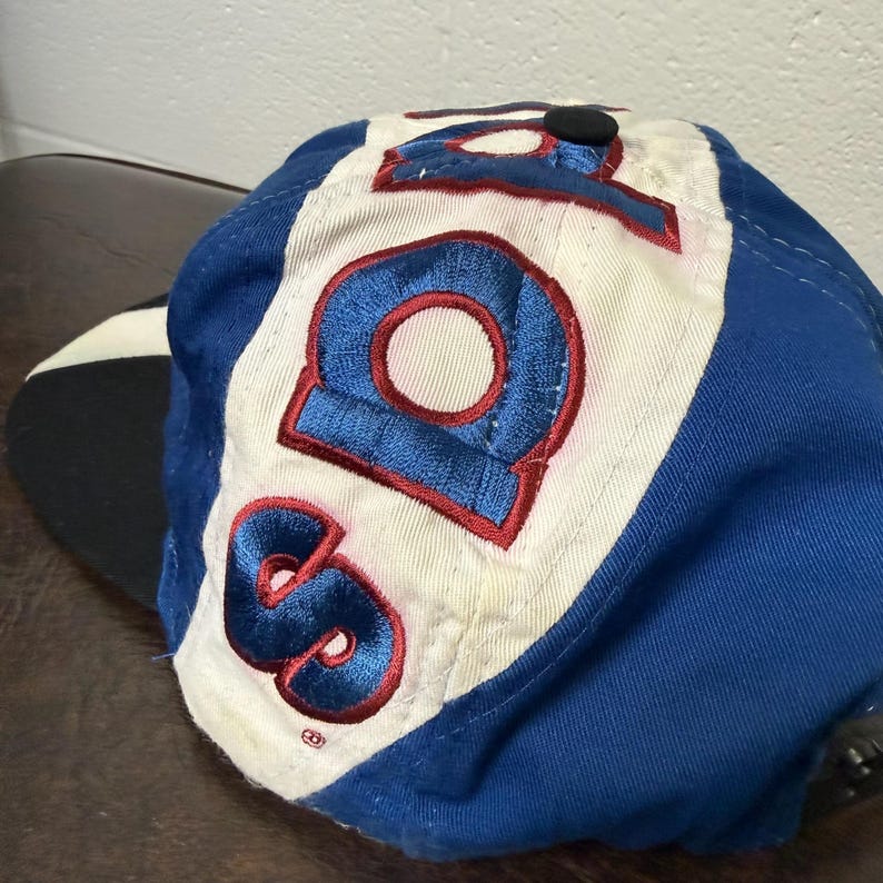May include: A blue and white baseball cap with the number "98" embroidered in blue with red trim. The cap has a black button on top and a black brim.