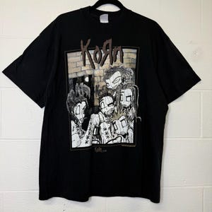 May include: Black t-shirt featuring a graphic of the band Korn. The design includes the band's name in brown lettering above a cartoon illustration of four figures. The shirt is hanging on a black hanger.