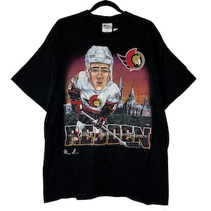 May include: Black t-shirt featuring a hockey player graphic with the name "REDDEN" in a stylized font. The graphic includes the Ottawa Senators logo and a depiction of a building. The shirt is hanging on a black hanger.