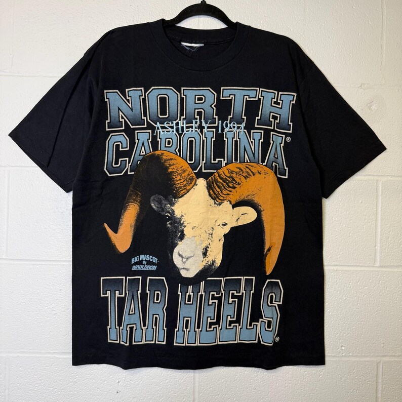 May include: Black t-shirt with the words "North Carolina" and "Tar Heels" in blue and white. It features a ram graphic with orange horns. The text "Asheville 1967" is also visible on the shirt.