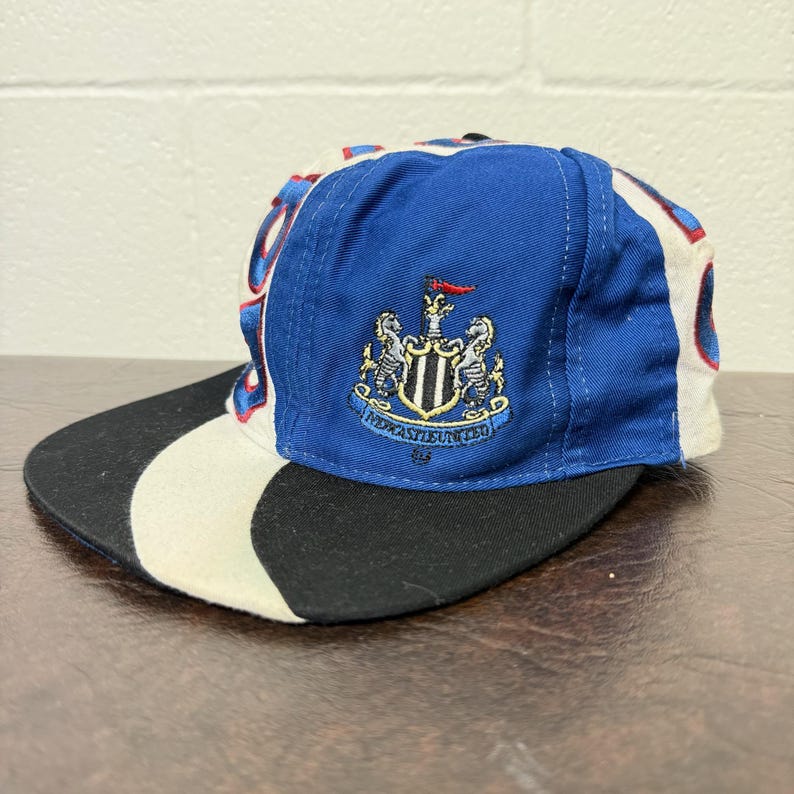 May include: A blue, white, and black baseball cap featuring the Newcastle United crest. The cap has a curved brim and a structured crown. The crest is embroidered on the front, with the team name below. The cap is made of a durable fabric.