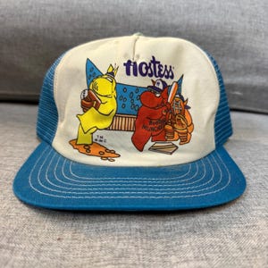 May include: A vintage Hostess baseball cap with a white front panel featuring cartoon characters and the Hostess logo in purple. The cap has a blue mesh back and a blue brim with white stitching. The design includes the text "Hostess" and "Hostess Munchies".