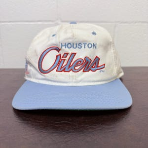 Op de afbeelding: Een vintage honkbalpet met een witte kroon en een lichtblauwe klep. De pet heeft de tekst "HOUSTON Oilers" in blauw script met een rode omlijning. Een klassiek sportaccessoire.