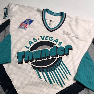 Może przedstawiać: Biała koszulka hokejowa z logo Las Vegas Thunder w kolorze turkusowym, czarnym i srebrnym. Koszulka ma numer "1" na lewym ramieniu i jest podpisana przez "Randy'ego Roenicka".