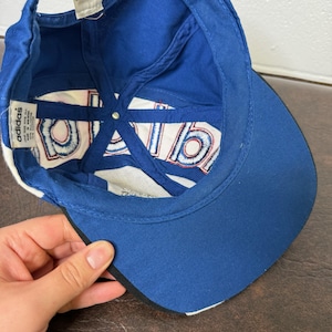 May include: A blue Adidas baseball cap with a white and red design on the underside. The cap has a curved brim and a white stripe along the edge. The Adidas logo is visible on a tag inside the cap.