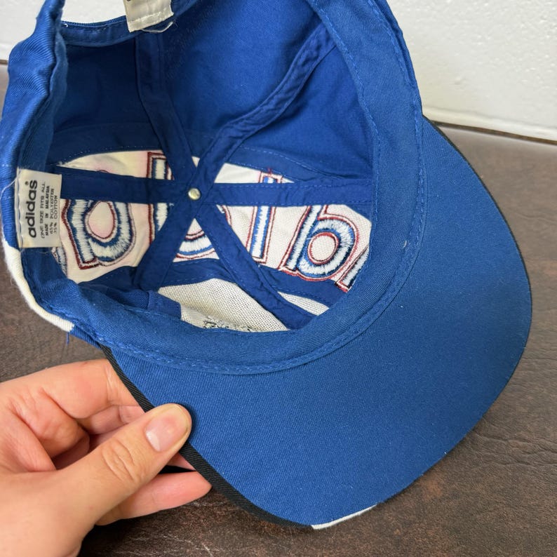 May include: A blue Adidas baseball cap with a white and red design on the underside. The cap has a curved brim and a white stripe along the edge. The Adidas logo is visible on a tag inside the cap.