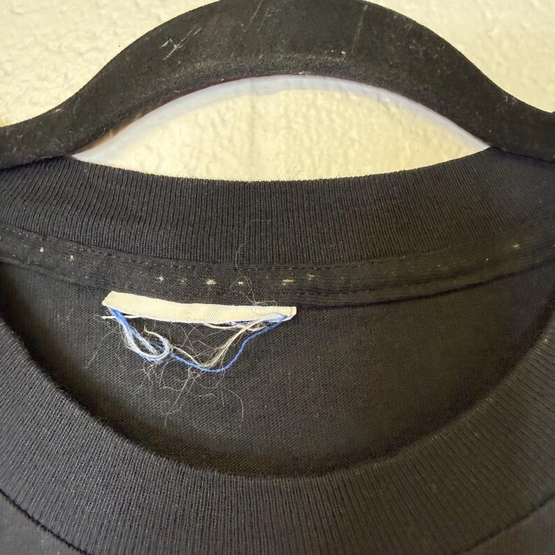 May include: Close-up of a black t-shirt neckline with a white label and blue thread. The shirt is hanging on a black hanger. The label is slightly frayed.