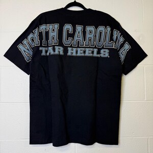 May include: Black t-shirt featuring the text "NORTH CAROLINA TAR HEELS" across the back in light blue and white. The shirt is displayed on a black hanger against a white brick wall.