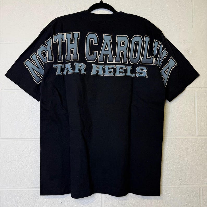 May include: Black t-shirt featuring the text "NORTH CAROLINA TAR HEELS" across the back in light blue and white. The shirt is displayed on a black hanger against a white brick wall.