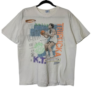 Może przedstawiać: Biała koszulka w stylu vintage z koszykarzem trzymającym piłkę, z napisem "Kelly Tripucka" i "Charlotte Hornets". Koszulka ma graficzny wzór z elementami i tekstem związanym z koszykówką.