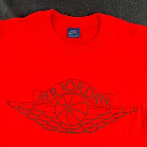 May include: Red t-shirt with a black Air Jordan logo featuring a basketball and wings. The shirt has a Nike tag with the size "Men's L" on the neck.