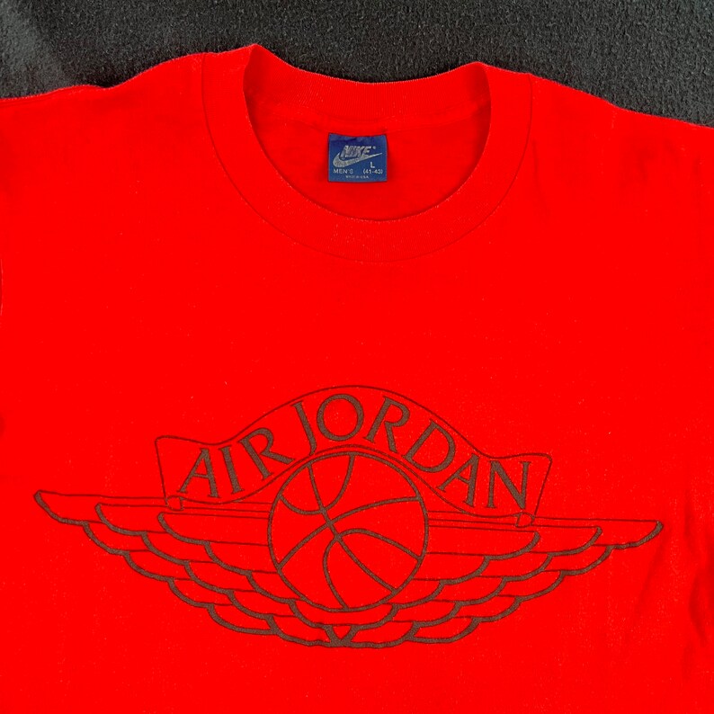 May include: Red t-shirt with a black Air Jordan logo featuring a basketball and wings. The shirt has a Nike tag with the size "Men's L" on the neck.