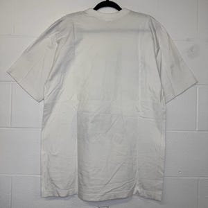 May include: A white, short-sleeved t-shirt hanging on a black hanger. The shirt is made of a soft, opaque fabric. The t-shirt is plain and has a crew neck.