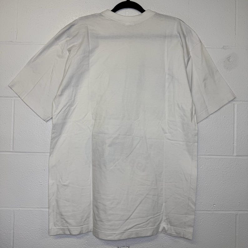 May include: A white, short-sleeved t-shirt hanging on a black hanger. The shirt is made of a soft, opaque fabric. The t-shirt is plain and has a crew neck.