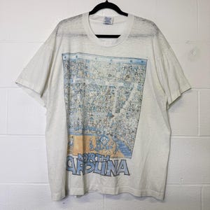 Puede incluir: Camiseta blanquecina con una ilustraci&oacute;n detallada de un partido de baloncesto y una multitud de personas en las gradas. Las palabras "North Carolina" est&aacute;n impresas en azul y gris en la parte inferior. La camiseta est&aacute; colgada de una percha negra.