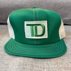 Peut inclure: Casquette de camionneur verte et blanche avec une visière plate. Le panneau avant est vert avec un écusson blanc brodé des lettres "TD" en vert. L'arrière en mesh est blanc et la visière est verte.