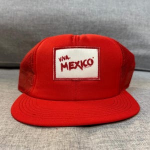 Peut inclure: Casquette de camionneur rouge avec un patch rectangulaire blanc portant l'inscription "VIVA MEXICO" en rouge. Le chapeau a un dos en filet et une visière incurvée. La casquette est de couleur rouge vif.