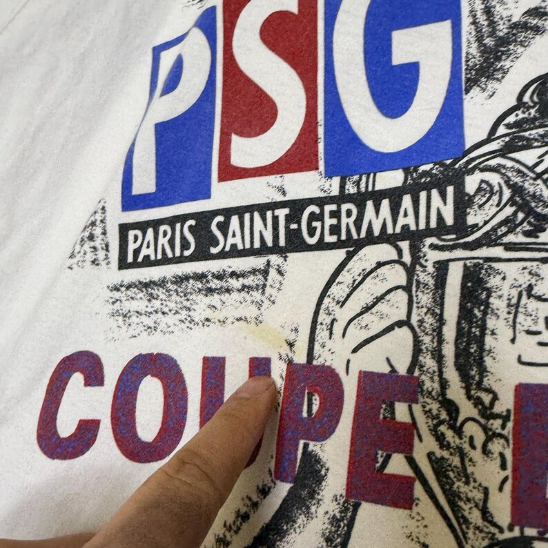 May include: White t-shirt with the text "PSG PARIS SAINT-GERMAIN COUPE". The letters "PSG" are in blue and red, with the words "PARIS SAINT-GERMAIN" in black. The word "COUPE" is in red and blue.