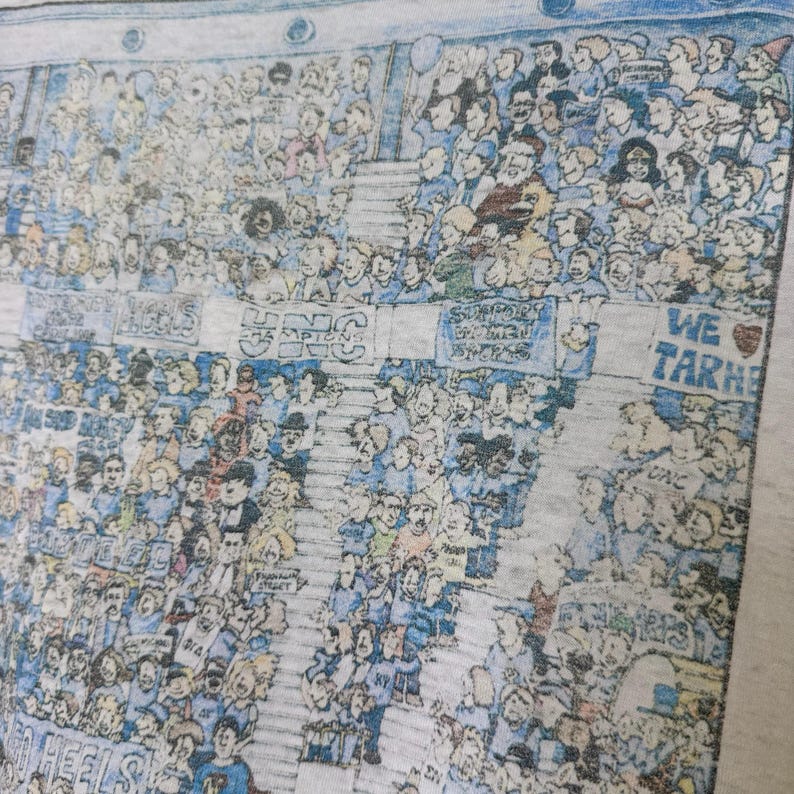 Puede incluir: Una camiseta gris claro con una ilustraci&oacute;n detallada de una multitud en un estadio. La obra de arte incluye numerosas figuras en azul y blanco, con pancartas y el texto "UNC Champions" y "We Tar Heels".