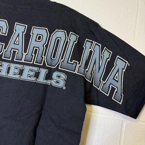 May include: Navy blue t-shirt featuring the text "CAROLINA HEELS" in light blue and white lettering. The design is on the upper back and sleeve. The shirt is made of a soft material.