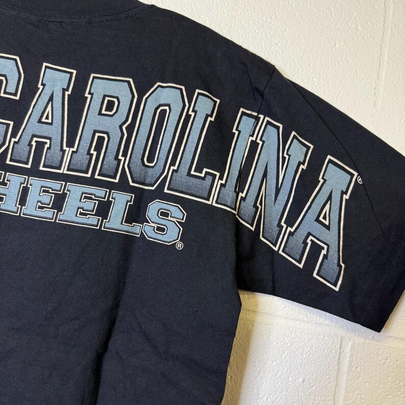 May include: Navy blue t-shirt featuring the text "CAROLINA HEELS" in light blue and white lettering. The design is on the upper back and sleeve. The shirt is made of a soft material.