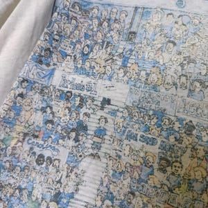 Puede incluir: Primer plano de una camiseta gris claro con una ilustraci&oacute;n detallada de una multitud de personas en azul y blanco. El texto incluye "Tar Heels #1" y "University of North Carolina".