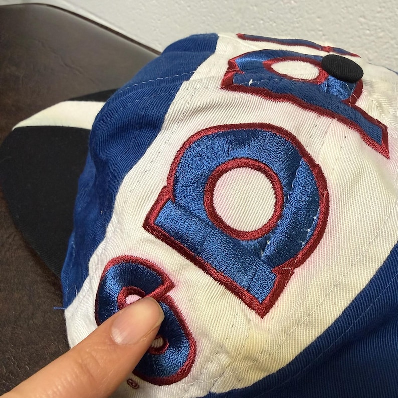 May include: A blue, white, and red baseball cap. The cap features the number "9" in blue with a red outline. The cap has a black button on top and a blue brim. The cap is made of a soft, possibly cotton, material.