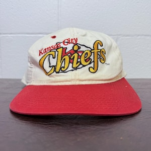 May include: A vintage baseball cap with a red bill and a cream-colored crown. The cap features the embroidered text "Kansas City Chiefs" in yellow and red, with a black outline. The hat is a classic sports accessory.