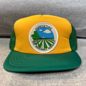 Peut inclure: Casquette de camionneur avec un panneau avant jaune et un dos et une visière en filet vert. Le chapeau présente un écusson circulaire avec les mots "AGRICULTURE OUR RENEWABLE RESOURCE" autour d'un graphique de champ, d'arbre et de ciel.