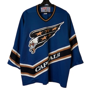 Puede incluir: Jersey de hockey azul con el logo de los Capitals de Washington. La camiseta tiene un cuello en V con ribete marrón, rayas blancas y marrones en las mangas y la palabra "CAPITALS" en la parte inferior.