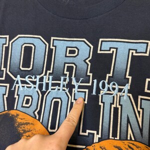 May include: Close-up of a navy blue t-shirt with the text "NORTH CAROLINA" in light blue and white lettering. The word "ASHLEY" and the number "1994" are also visible in light blue.