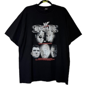 Puede incluir: Camiseta negra con el texto "King of the Ring" y el logo de la WWF. La camiseta muestra imágenes de luchadores. También se ve el texto "Federation Championship Match" y "Taking Control of The World Wrestling Federation".