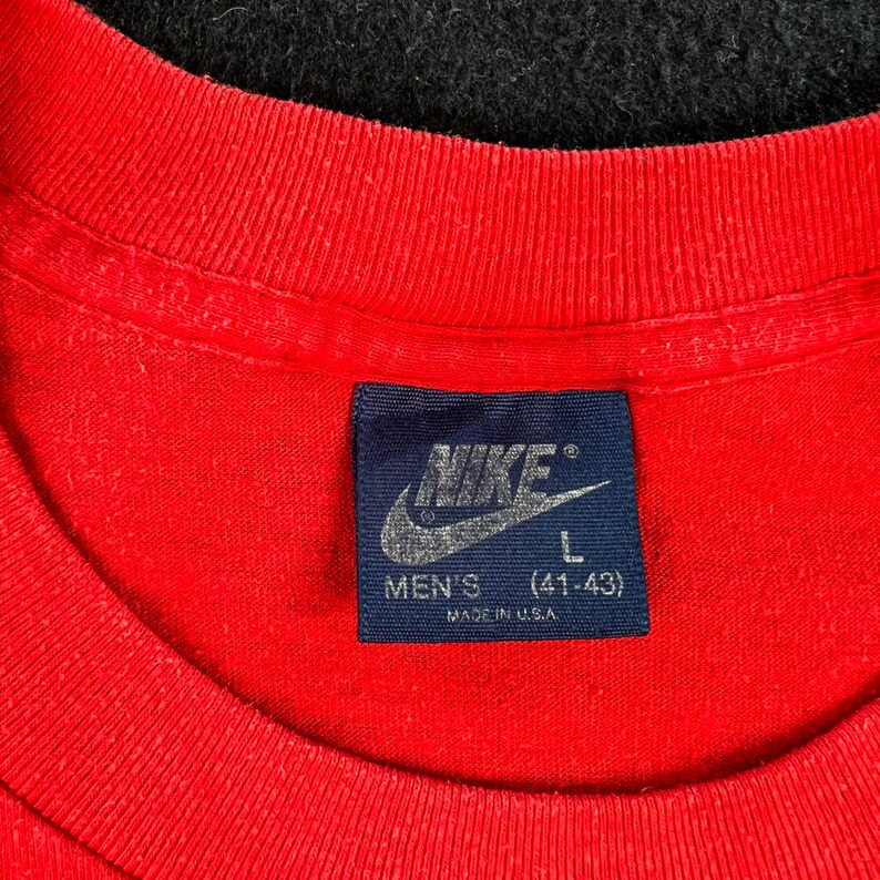 May include: A red t-shirt with a blue Nike tag that says "Nike" in white lettering and "Men's (41-43) Made in U.S.A."