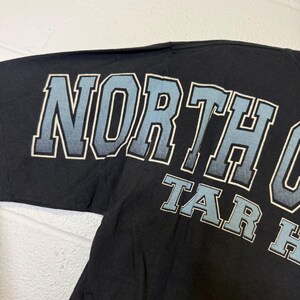 May include: Black t-shirt featuring the text "NORTH CAROLINA" and "TAR HEELS" in light blue and white. The text is in a bold, collegiate-style font.