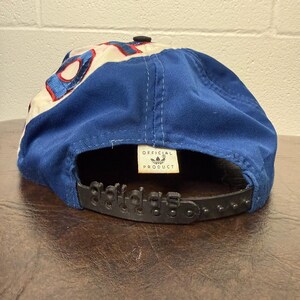 May include: Blue and white baseball cap with the Adidas logo on the back. The cap has a black adjustable strap and a small tag that says "OFFICIAL PRODUCT". The cap is made of a durable fabric.