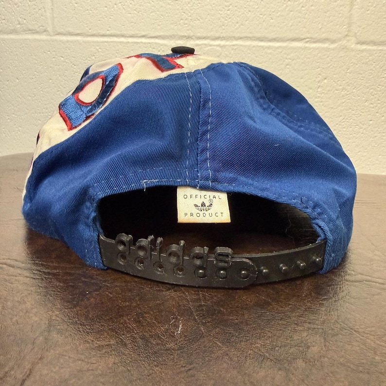 May include: Blue and white baseball cap with the Adidas logo on the back. The cap has a black adjustable strap and a small tag that says "OFFICIAL PRODUCT". The cap is made of a durable fabric.