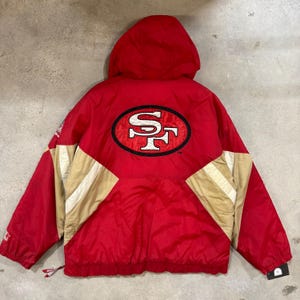 May include: Red and gold hooded jacket with the San Francisco 49ers logo on the back. The jacket has a red body with gold and white accents on the sleeves and shoulders. The jacket has a Starter logo on the left sleeve.