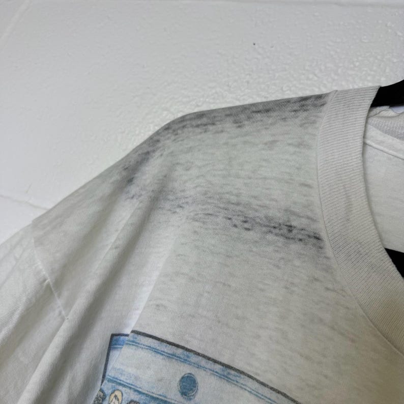 Puede incluir: Primer plano de una camiseta blanca con un dise&ntilde;o descolorido. El &aacute;rea del hombro de la camiseta muestra signos de desgaste y decoloraci&oacute;n. El cuello es blanco liso y la tela parece ser una mezcla de algod&oacute;n ligero. La camiseta est&aacute; colgada en una percha negra.