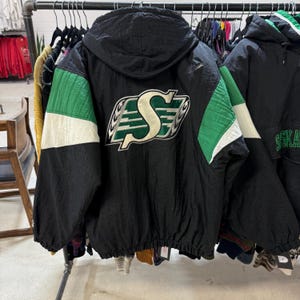 Puede incluir: Chaqueta negra con capucha y detalles en verde y blanco. Cuenta con un gran logotipo bordado en la espalda. La chaqueta está colgada en una percha con otras prendas.