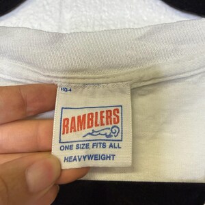 Puede incluir: Primer plano de un cuello de camiseta blanca con una etiqueta. La etiqueta tiene un borde azul y presenta la palabra "RAMBLERS" en rojo, junto con el texto "ONE SIZE FITS ALL" y "HEAVYWEIGHT".