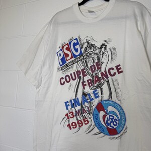 May include: White t-shirt featuring a graphic of a trophy and text. The text includes "PSG PARIS SAINT-GERMAIN", "COUPE DE FRANCE FINALE 13 MAI 1995", and a Racing Club de Strasbourg logo.