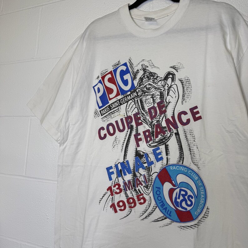 May include: White t-shirt featuring a graphic of a trophy and text. The text includes "PSG PARIS SAINT-GERMAIN", "COUPE DE FRANCE FINALE 13 MAI 1995", and a Racing Club de Strasbourg logo.