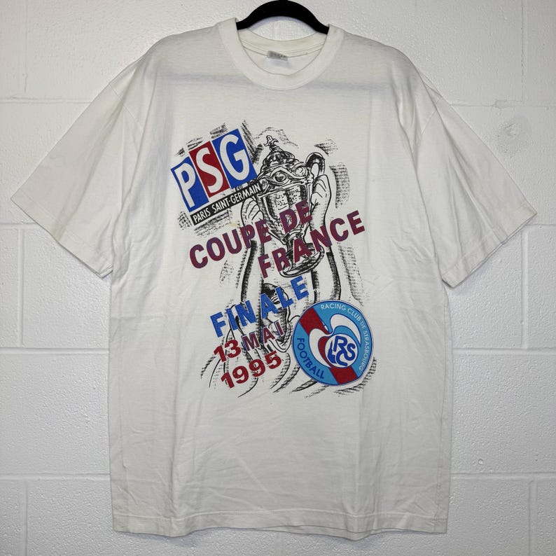 May include: White t-shirt featuring a graphic design commemorating the Coupe de France final. The design includes the PSG logo, the words "COUPE DE FRANCE FINALE 13 MAI 1995", and the Racing Club de Strasbourg logo. The shirt is short-sleeved.