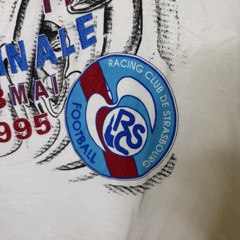 May include: White t-shirt featuring a Racing Club de Strasbourg logo in a circular design with blue, red, and white colors. The shirt also has text that reads "FINALE 3 MAI 1995".