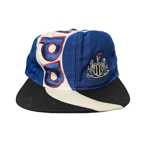 May include: A blue, white, and black baseball cap with the letters "NU" in red and white. The cap features a team crest on the side and a black brim. The hat is a vintage style.