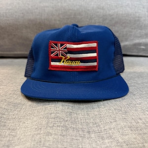 May include: A blue trucker hat with a curved brim and mesh back. The front features a patch with the Hawaiian flag and the word "Hawaii" in gold script. The hat is a classic style, suitable for casual wear.
