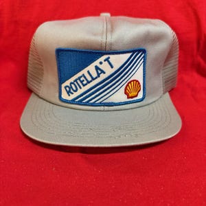 May include: A gray trucker hat with a mesh back and a curved brim. The front features a rectangular patch with the text "ROTELLA T", blue stripes, and a Shell logo. The hat is set against a red background.