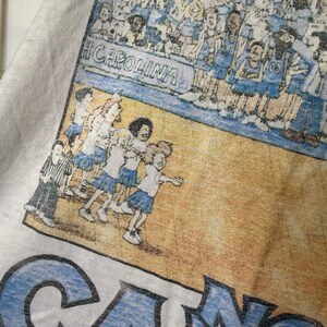 Puede incluir: Camiseta blanca con una ilustraci&oacute;n vintage de baloncesto. El dise&ntilde;o incluye el texto "H CAROLINA" y figuras de dibujos animados de animadoras y jugadores en azul y blanco. La camiseta tiene un aspecto retro descolorido.