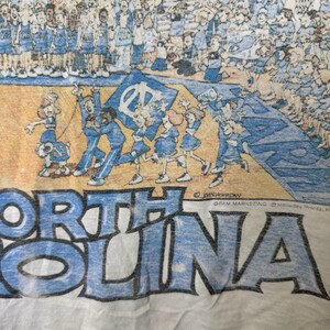 Puede incluir: Camiseta blanca con una ilustraci&oacute;n de dibujos animados de un equipo de baloncesto y animadoras. Las palabras "NORTH CAROLINA" est&aacute;n impresas en letras grandes de color azul claro en la parte inferior. La ilustraci&oacute;n incluye un signo de la paz.